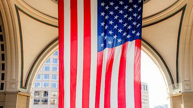A large American flag hangs between arched columns of a circular, open-air building, with a city street and trees below.
