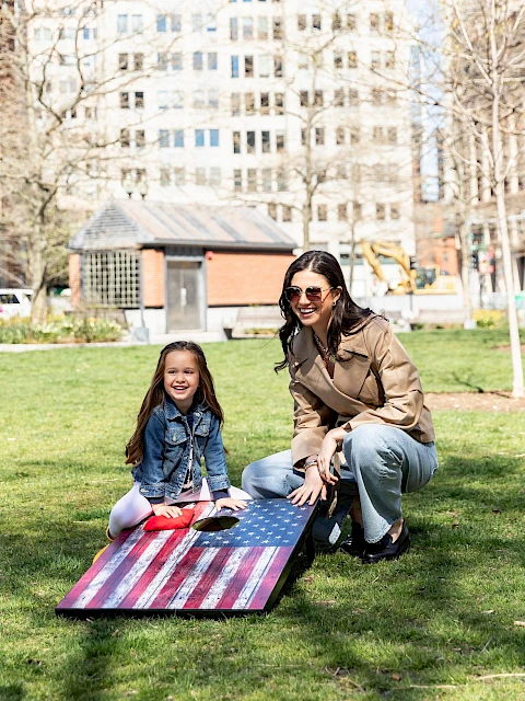 Three people enjoy a sunny day in a city park, with a girl and woman on a striped blanket while a child runs nearby.