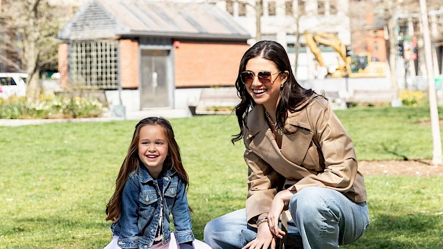 Three people enjoy a sunny day in a city park, with a girl and woman on a striped blanket while a child runs nearby.