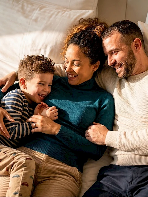 A joyful family lies together on a bed, parents smiling as they hug their child, a cozy, loving moment captured at home.