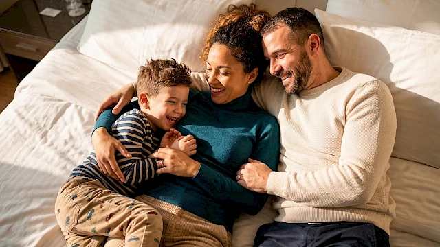 A joyful family lies together on a bed, parents smiling as they hug their child, a cozy, loving moment captured at home.