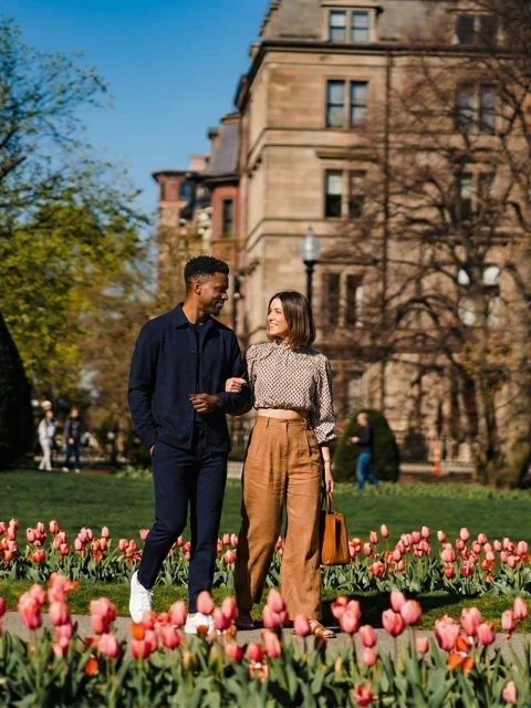 A couple walks hand-in-hand through a tulip garden in front of a brick-building, spring sun, green trees, and neatly trimmed bushes in bloom.