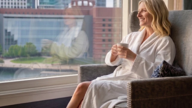 A woman in a white robe sits by a window, sipping coffee and enjoying the city view from a cozy hotel lounge.