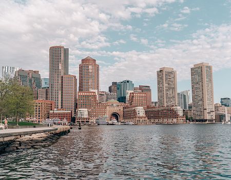 Boston skyline along the harbor with tall buildings, calm water in the foreground, a few boats, and a partly cloudy sky.