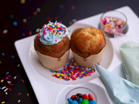 Two cupcakes with sprinkles, candy, and a piping bag on a white plate