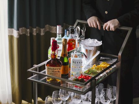 A person stands behind a cart with assorted liquor bottles, glasses, ice, and