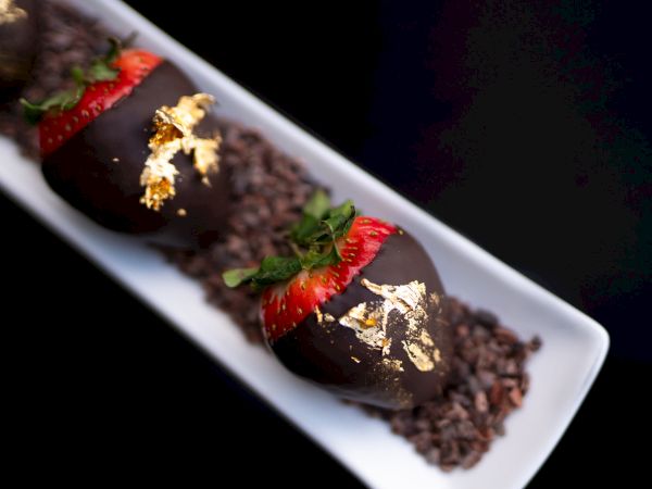 Three chocolate-covered desserts with strawberries and gold leaf on a plate, presented elegantly