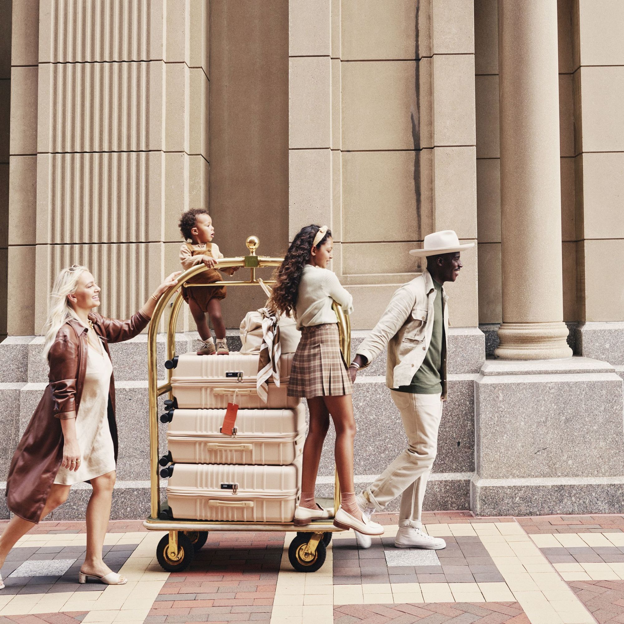 A group of four people, two women and two men, walk past a grand, stone fa&ccedil;ade with tall columns. They push a luggage cart across a checkered pavement. The scene looks like a travel moment.
