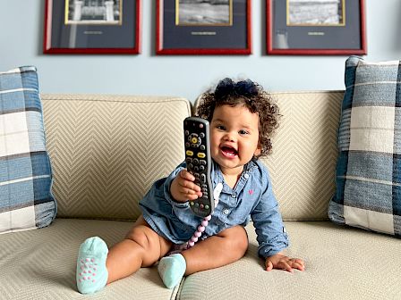 A baby sits on a sofa holding a phone, six framed black-and-white photos hanging on a light blue wall above, cheerful mood.