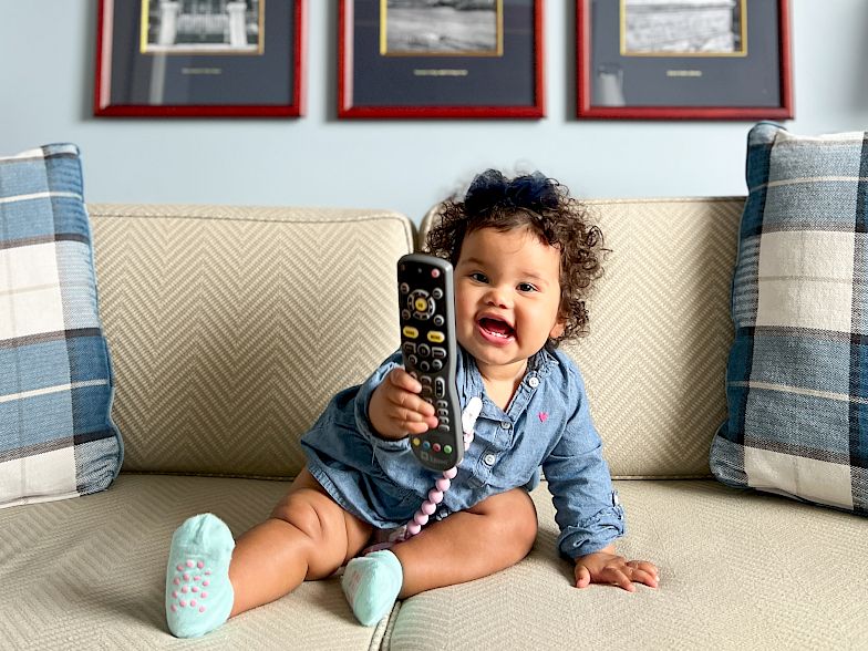 A baby sits on a sofa holding a phone, six framed black-and-white photos hanging on a light blue wall above, cheerful mood.