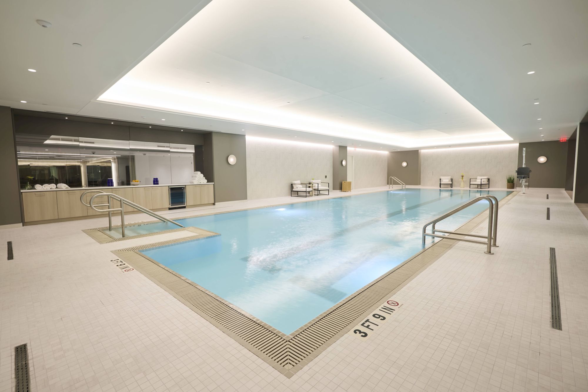 An indoor modern swimming pool area with clean white tiles, blue water, lounge chairs, and a reception/kitchen area in the background, well lit and quiet.