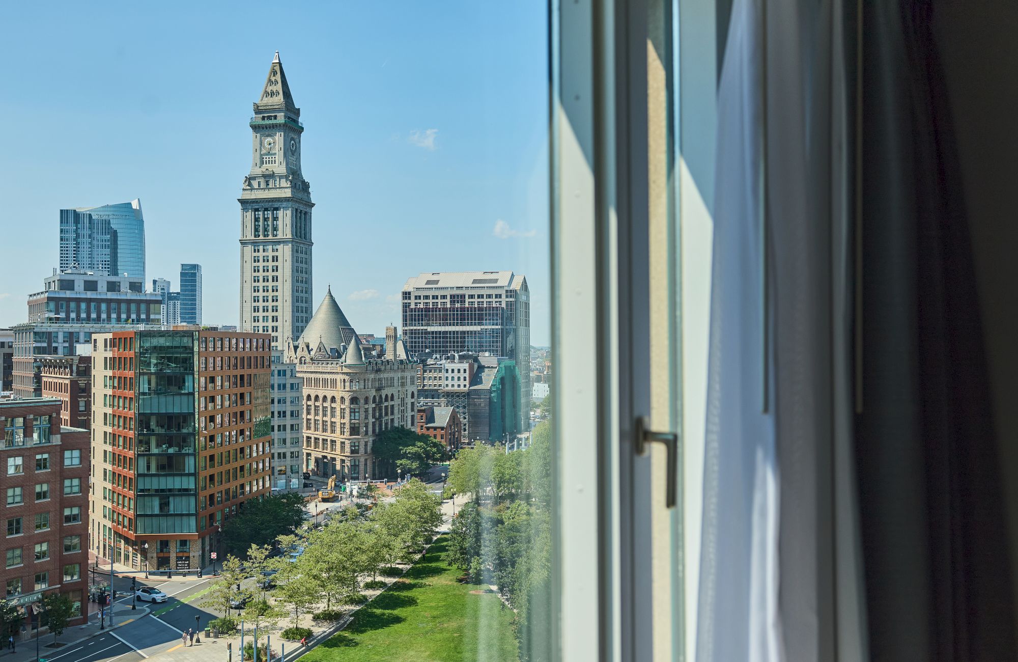 City skyline with tall clock tower, modern buildings, a green park along a street, viewed from a window with curtains.