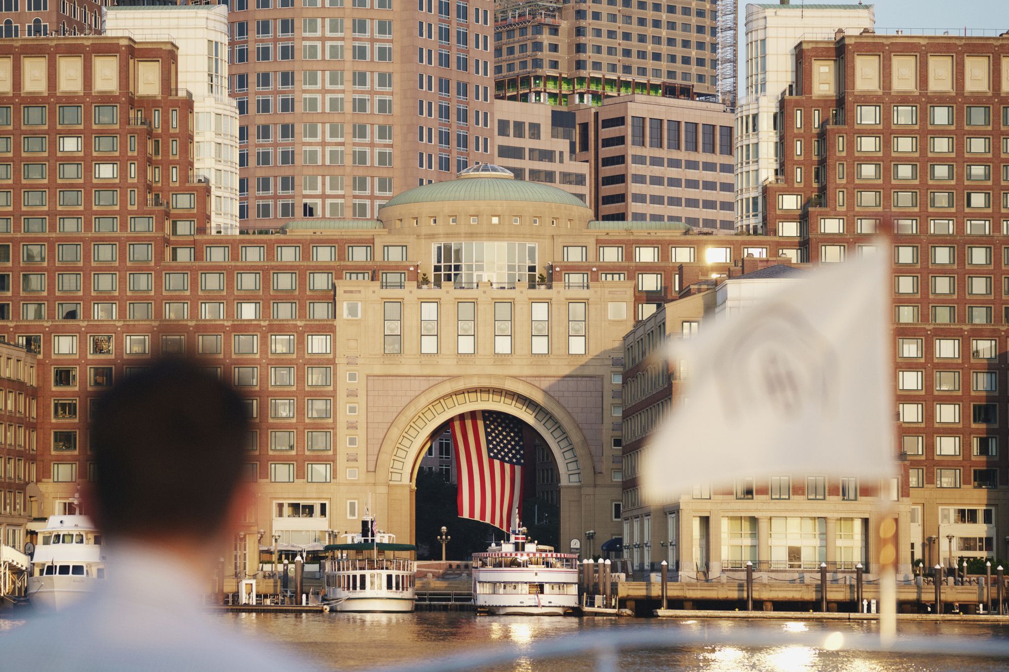 A waterfront cityscape with brick buildings, an arched entrance, boats, and a flag waving in front of a crowd near the water.