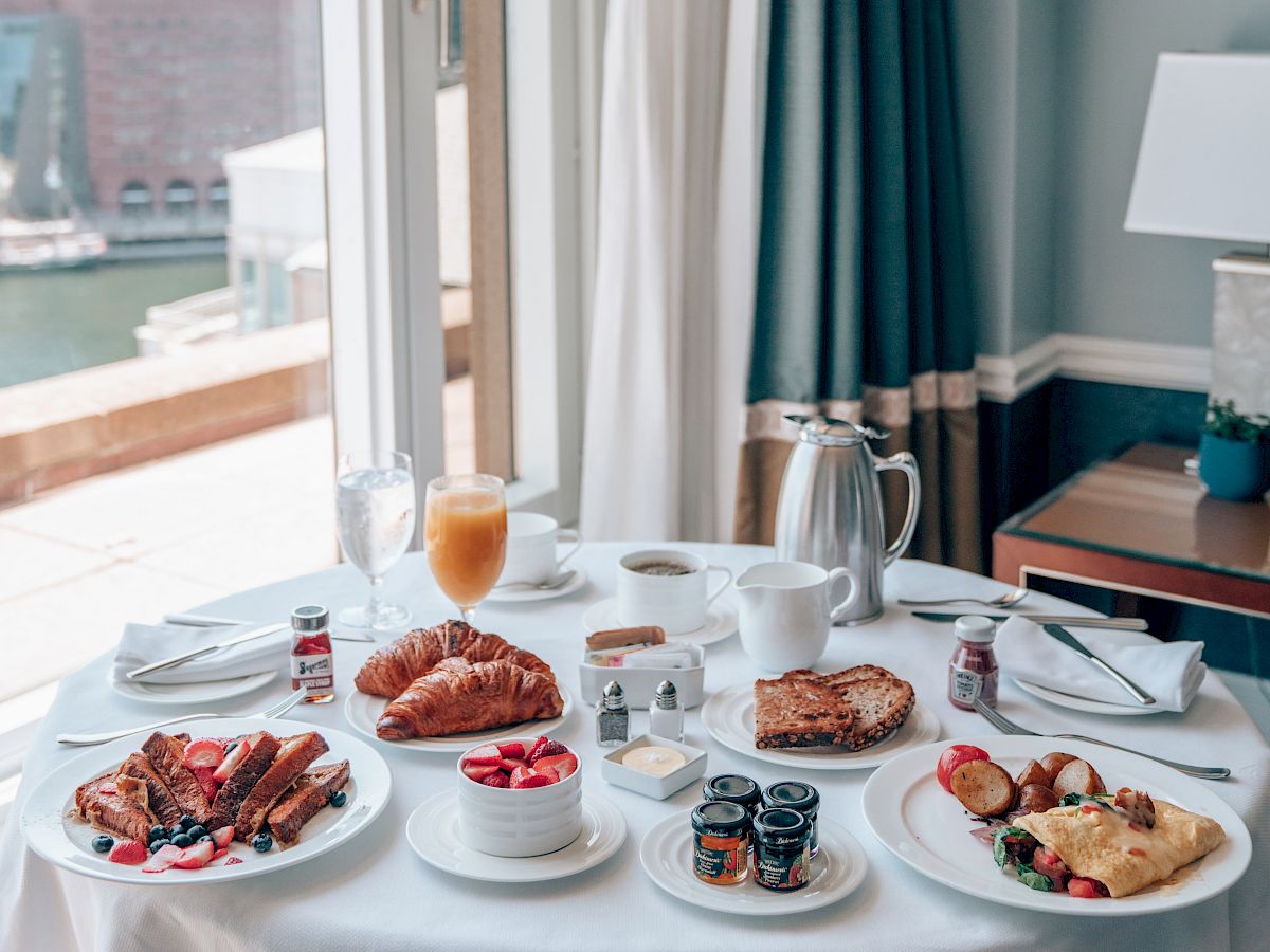 A bright hotel room breakfast spread: croissants, berries, waffles, yogurt, coffee, juice, and pastries on a white table by a sunny window.