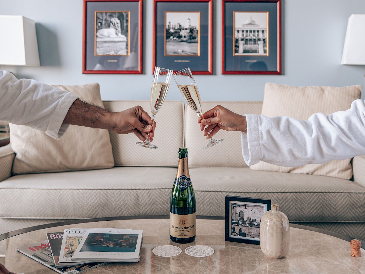 Two people toast with champagne in a modern living room, framed photos on the wall, a marble coffee table, and a bottle hovering mid-toast.