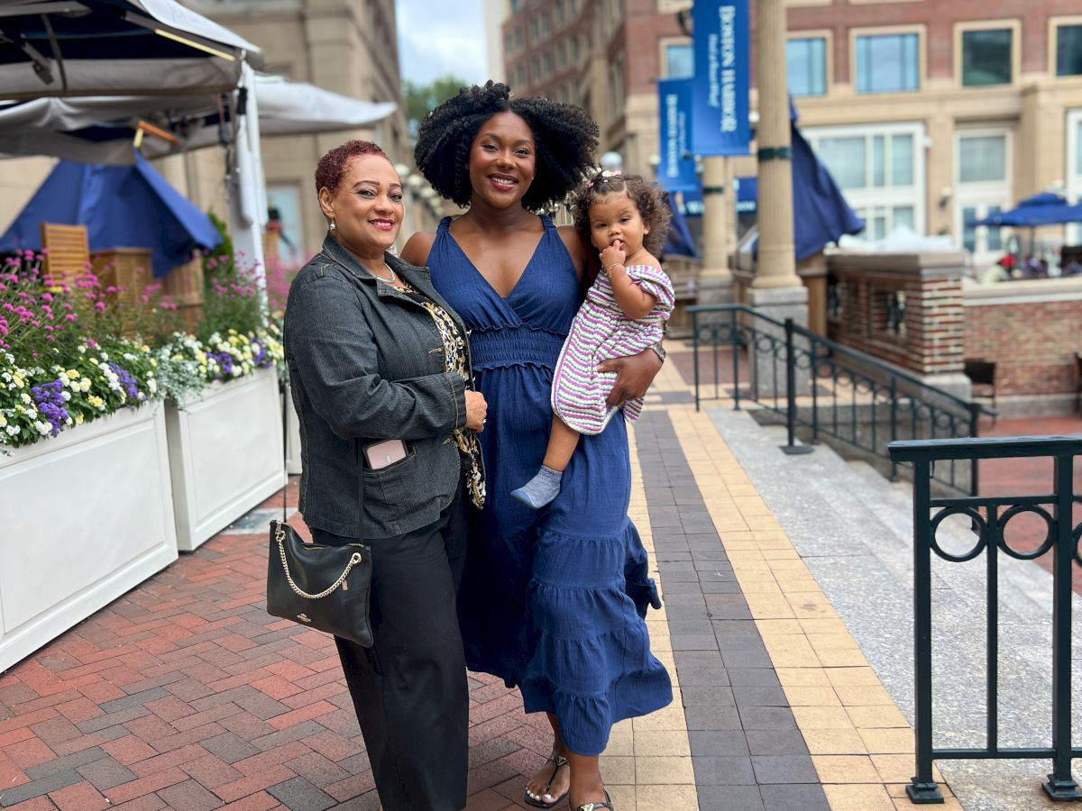 Three women posing on a brick sidewalk in an urban plaza; two adults with a child, smiling and dressed nicely.