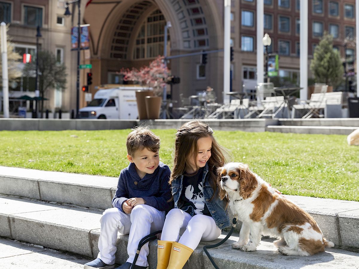 Two kids sit on steps with a friendly dog in a city park, modern buildings and an arched gateway behind them, sunny day.