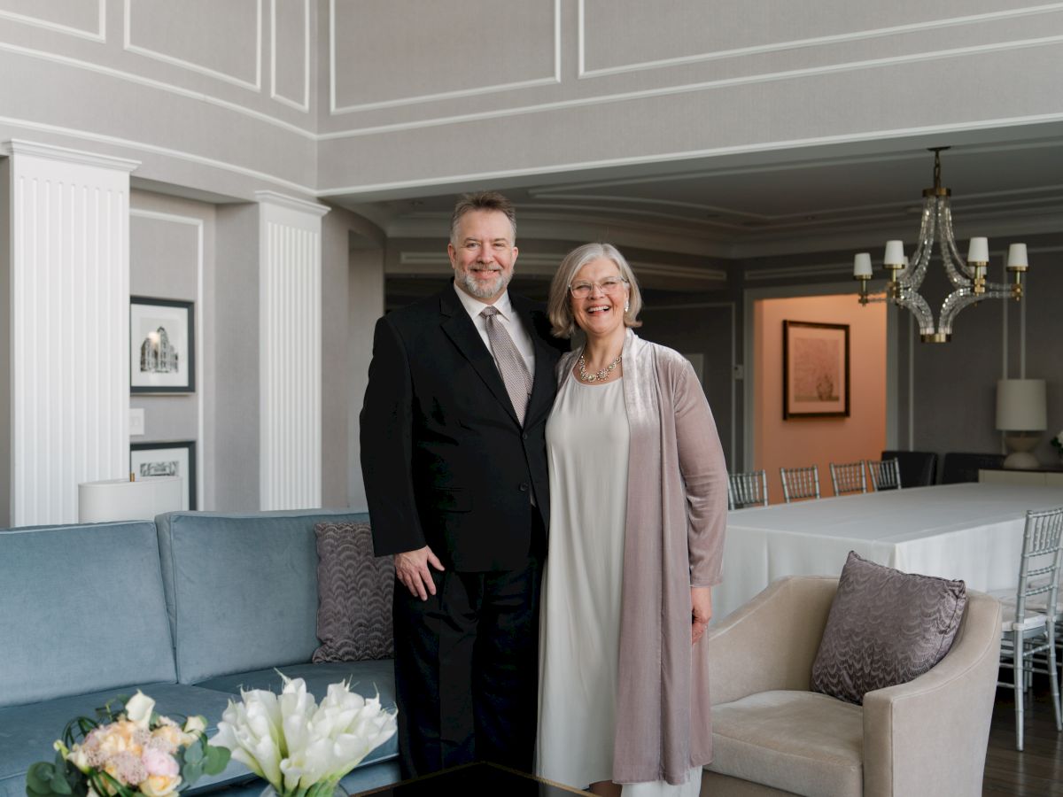 A well-dressed couple stands in a stylish, modern living room with blue accents, a glass coffee table, and floral arrangements on top.