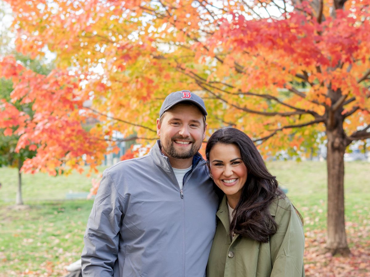 A couple posing outdoors in fall foliage, smiling beside a path with vibrant orange and red trees behind them.
