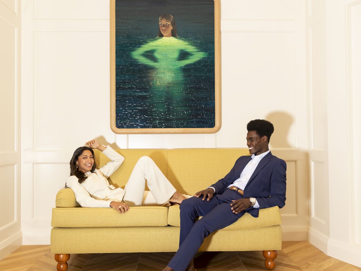 Two stylishly dressed people lounge on a yellow sofa in a bright room, with a framed green swimmer image above them on the wall.