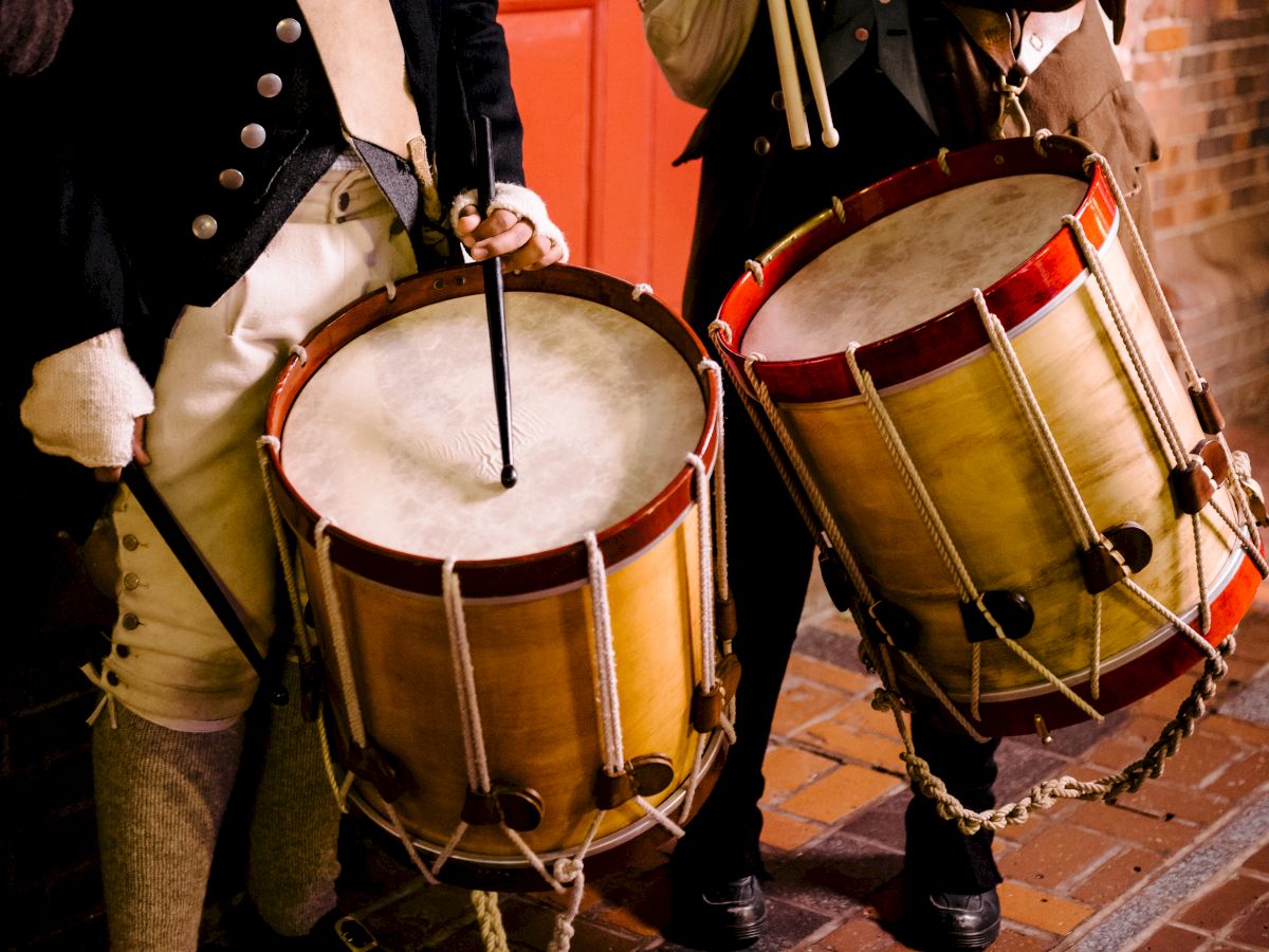 Two people playing marching drums with wooden shells, drumsticks raised, wearing period outfits; brick wall background.