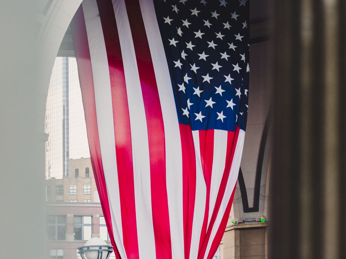 An American flag hangs between buildings, seen through a window arch, fluttering above a city street.