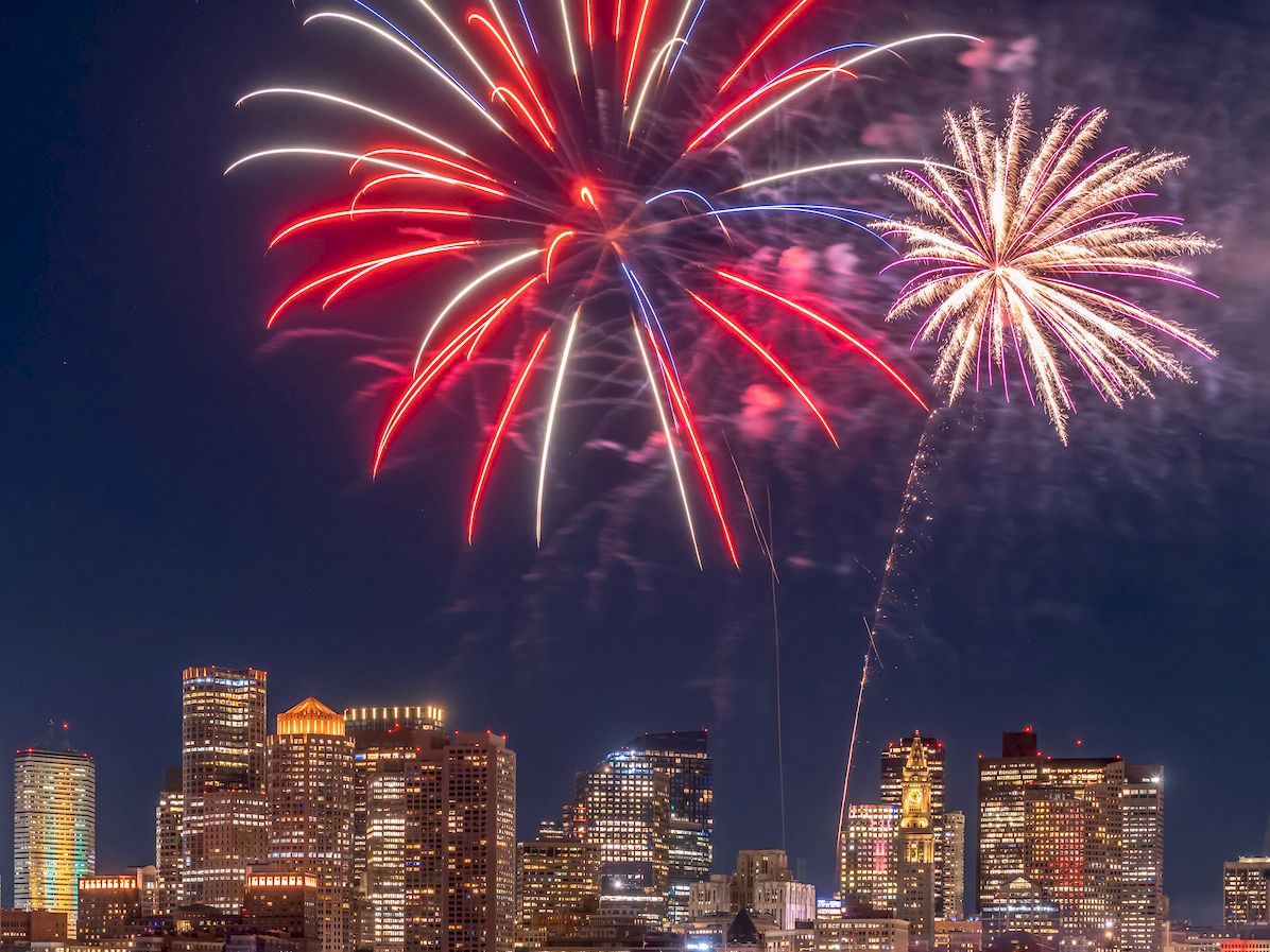 Colorful fireworks over a city skyline at night, reflected on the water, with tall buildings glowing.
