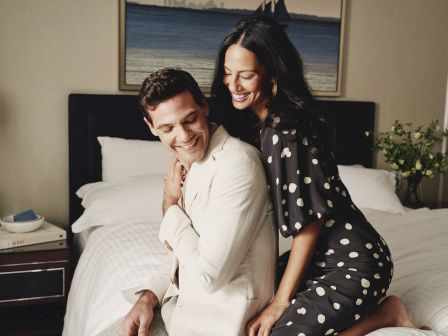 A couple sits on a bed indoors, smiling and embracing; the man in a light suit and the woman in a black polka-dot dress, cozy hotel-like room.