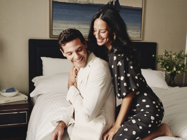 A couple sits on a bed indoors, smiling and embracing; the man in a light suit and the woman in a black polka-dot dress, cozy hotel-like room.