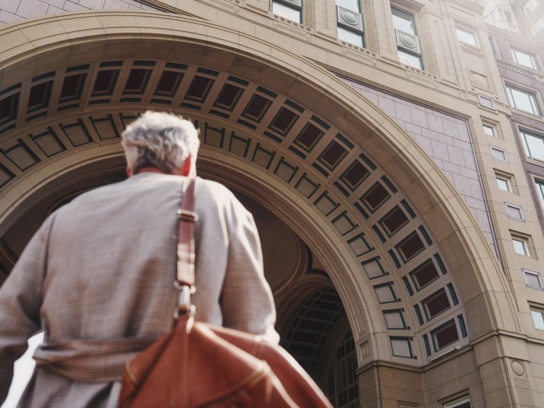 A man with a tan coat and brown bag is entering a grand arched building entrance.