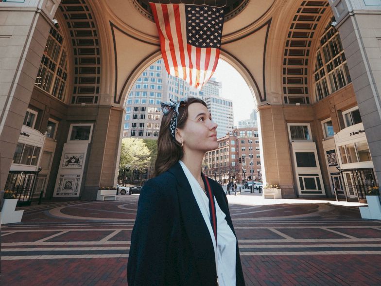 A young woman stands under a large arched gateway with an American flag hanging above, in a brick plaza setting.