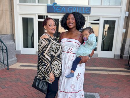 Three people stand outside a building entrance; two women smiling, one holding a child, all dressed nicely for a casual group photo.