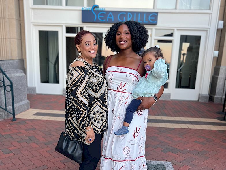 Three people stand outside a building entrance; two women smiling, one holding a child, all dressed nicely for a casual group photo.