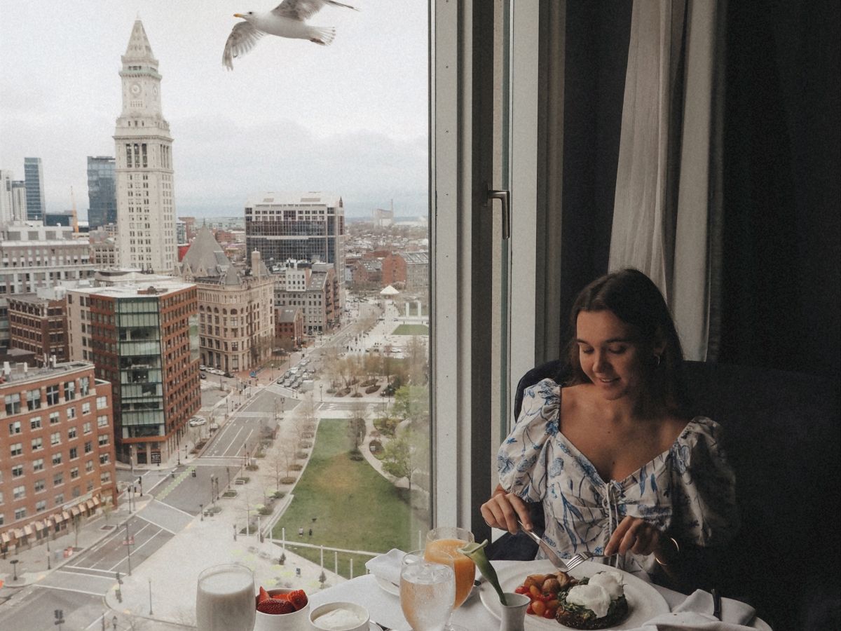 A woman dines at a high-rise window table with a city view, birds flying outside, and a lavish breakfast spread including pastries, fruit, and coffee.