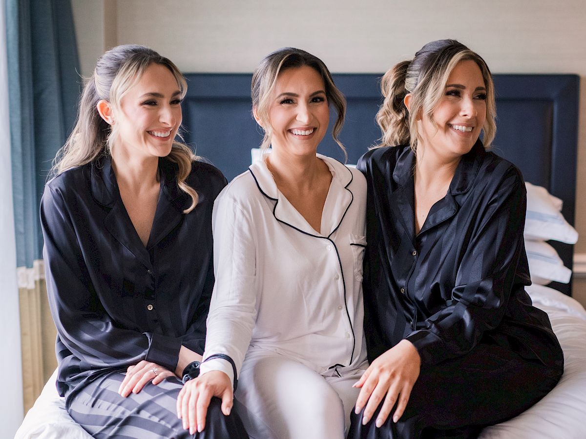 Three women sit on a bed in matching satin pajamas, smiling; two wear black and one white, slippers on a cozy hotel room floor.