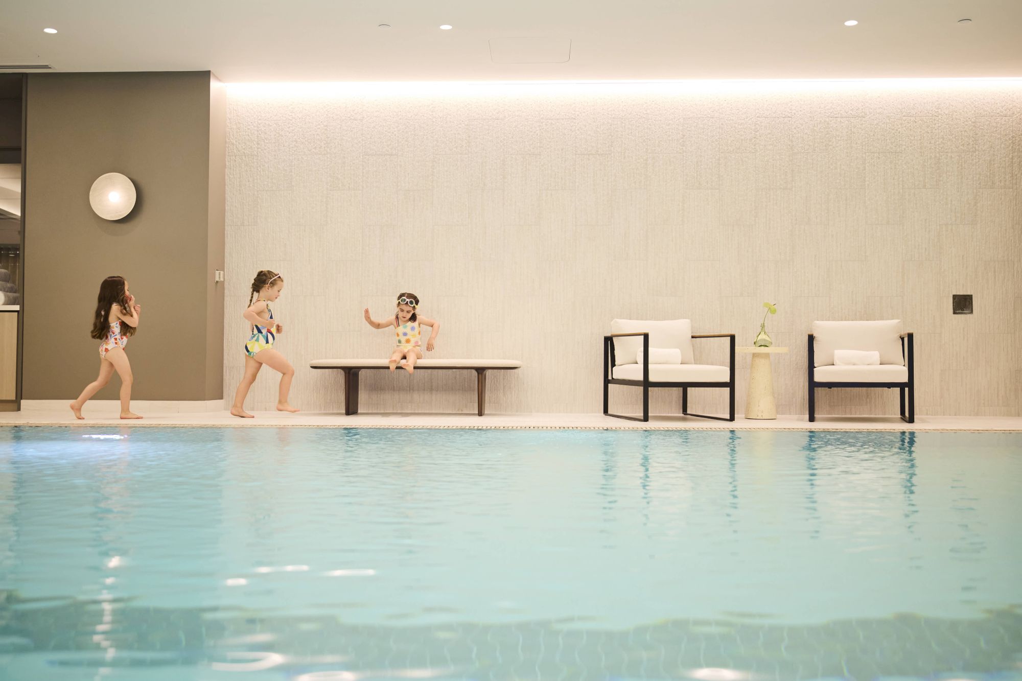 Kids play by an indoor pool: two girls running, one at the table, another in a chair area; calm, bright lounge with two white chairs.