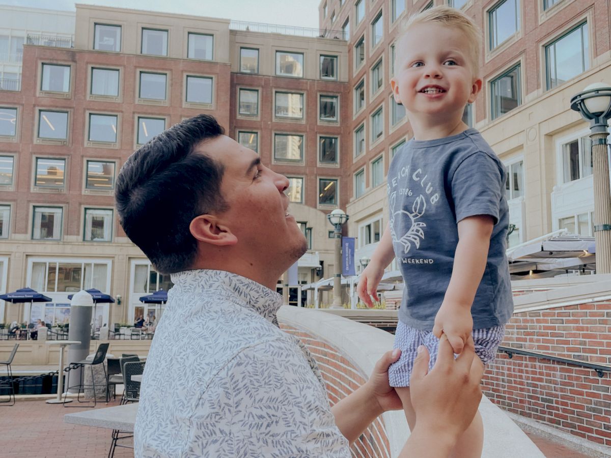 A man lifts a smiling toddler near a brick plaza with tall buildings in the background, capturing a joyful moment between them.