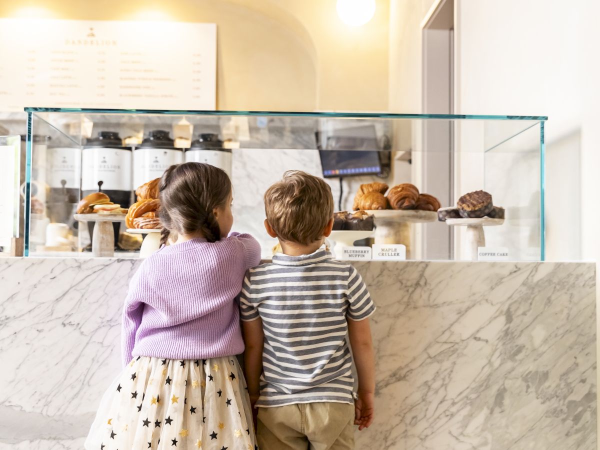 Two kids stand at a marble cafe counter, looking at pastries behind a glass display; girl wears a purple sweater and polka-dot skirt, boy in striped shirt and beige pants. End sentence.