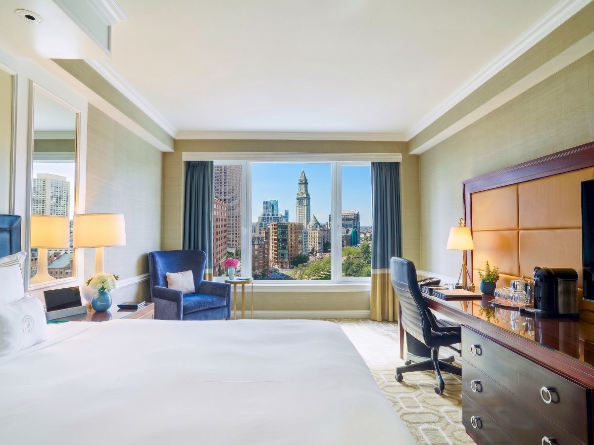 Bright hotel room with a bed, desk, chairs, lamps, and a window view of a city skyline.