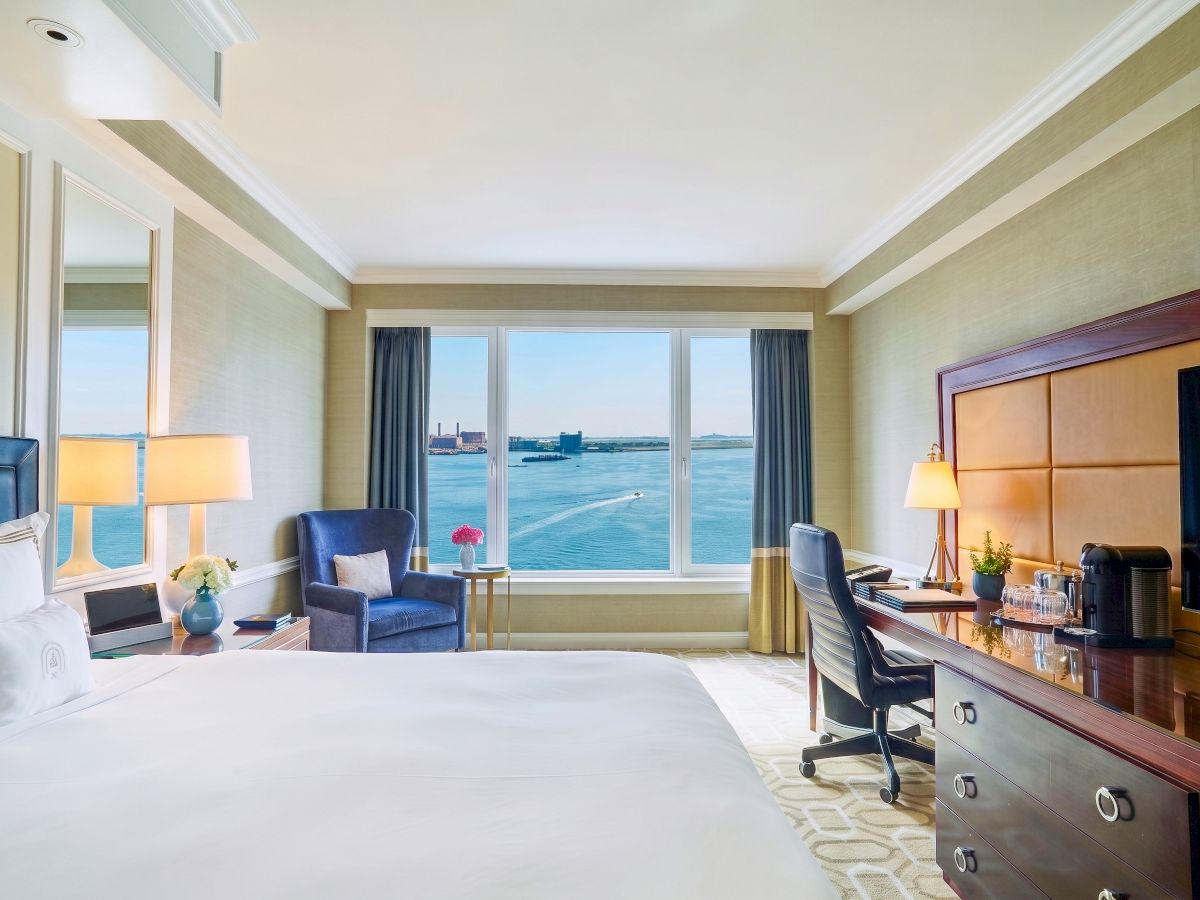 A bright hotel room with a bed, lounge chairs, desk and coffee setup, plus large windows overlooking a harbor and ocean view.