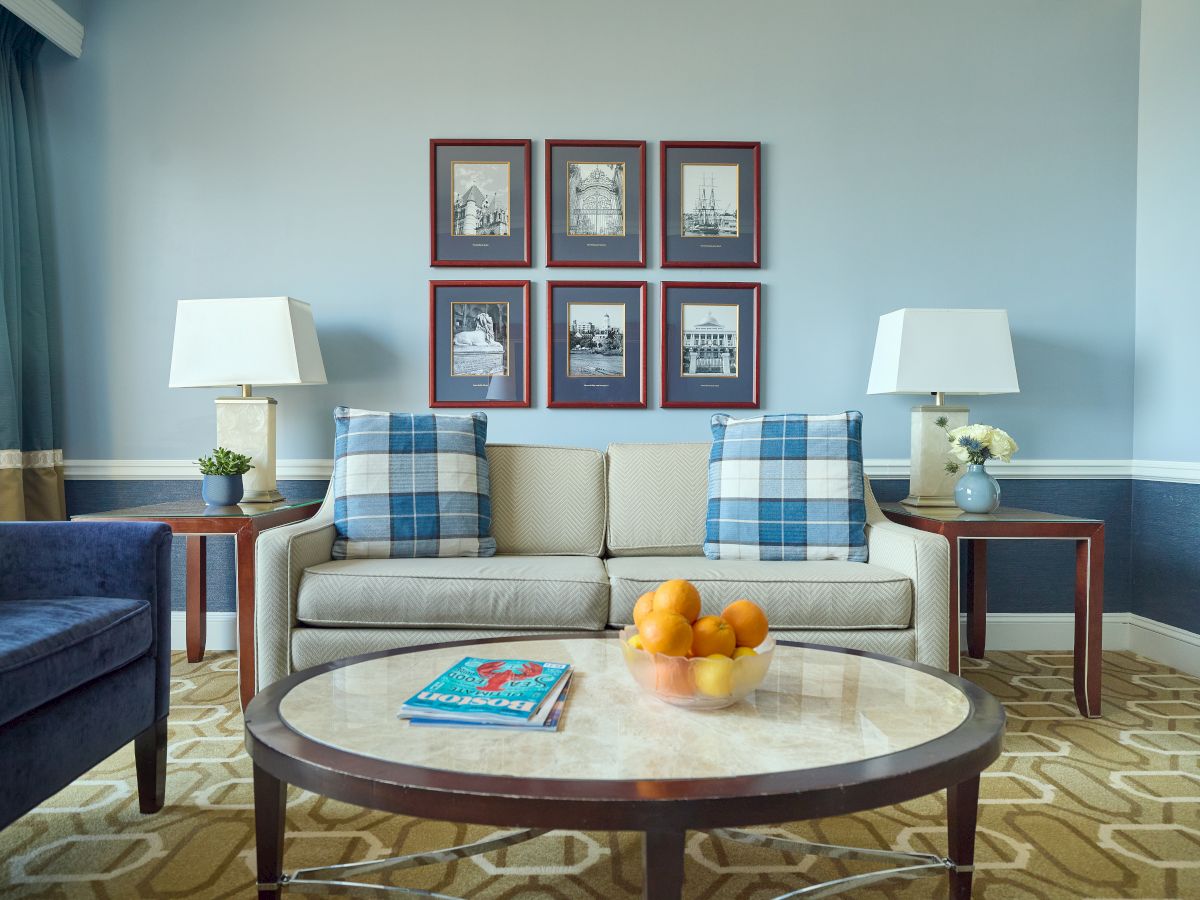 A cozy living room with a sofa, plaid pillows, two lamps, framed maritime artwork, and a coffee table with fruit and a book.