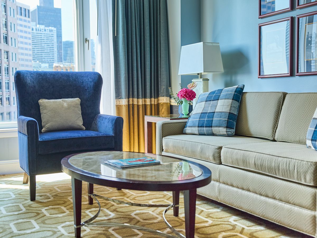A modern living room with a blue armchair and couch, a lamp, floral decor, a round coffee table, and tall curtains overlooking a city.