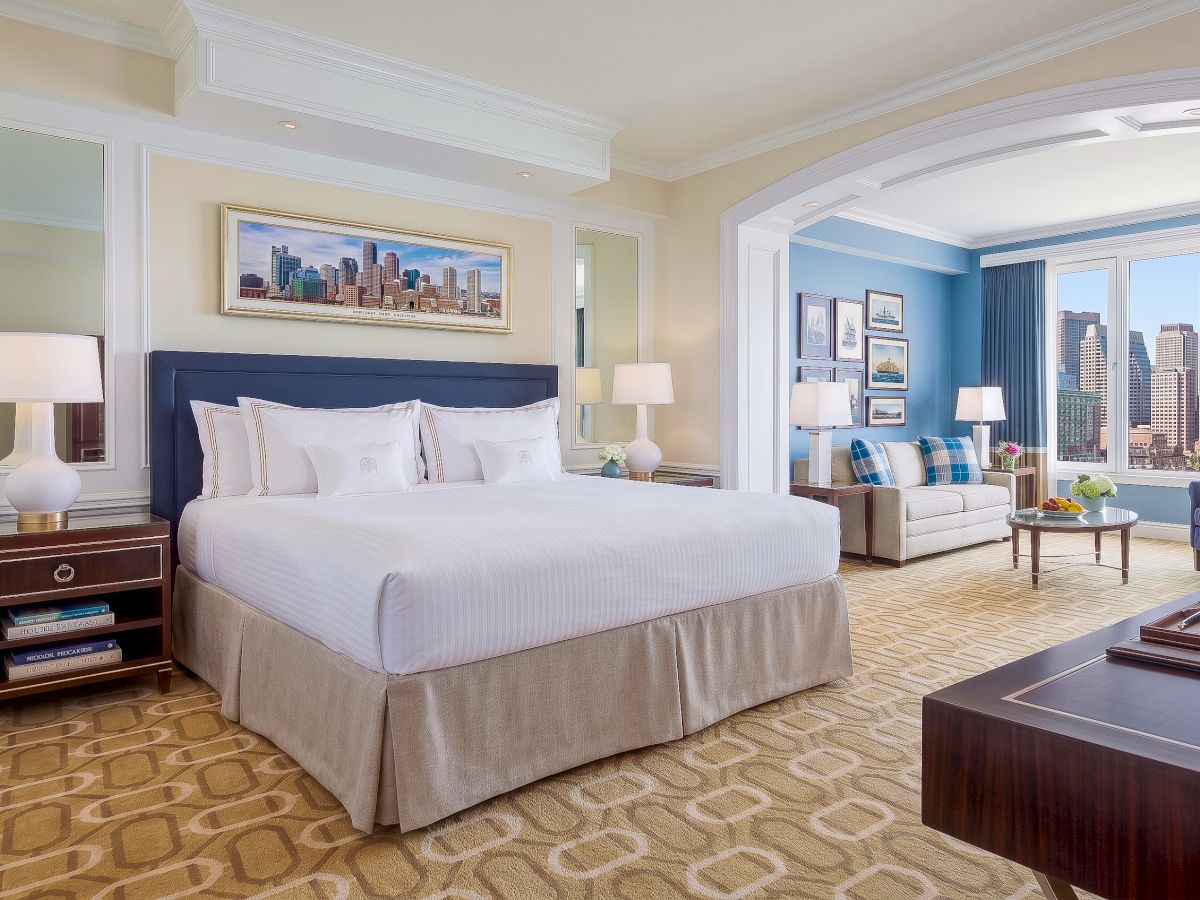 Luxury hotel bedroom with a king bed, lamps and lounge, plus large windows showing a city skyline view.