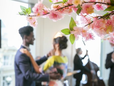 A couple dancing at a stylish indoor party with musicians in the background and pink blossoms in the foreground.