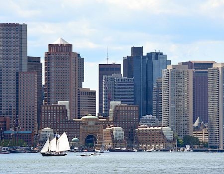 A city skyline by the water with tall buildings and a few sailboats near the shore, a bustling downtown against a cloudy sky.