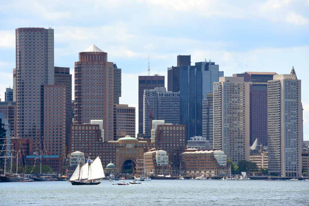 A city skyline by the water with tall buildings and a few sailboats near the shore, a bustling downtown against a cloudy sky.