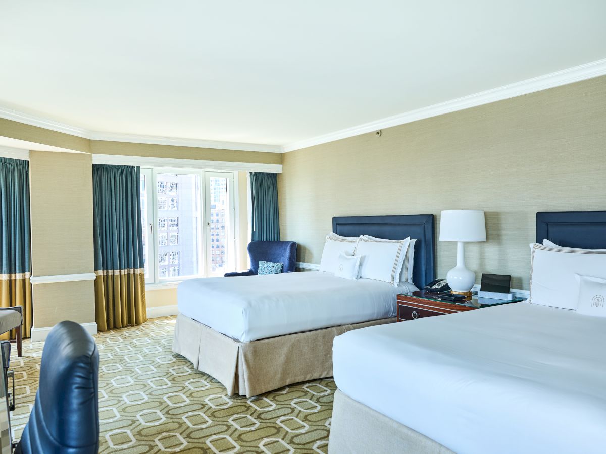 Two queen beds in a hotel room with nightstands, lamps, a desk, a chair, and windows with blue curtains; neutral tones and patterned carpet.