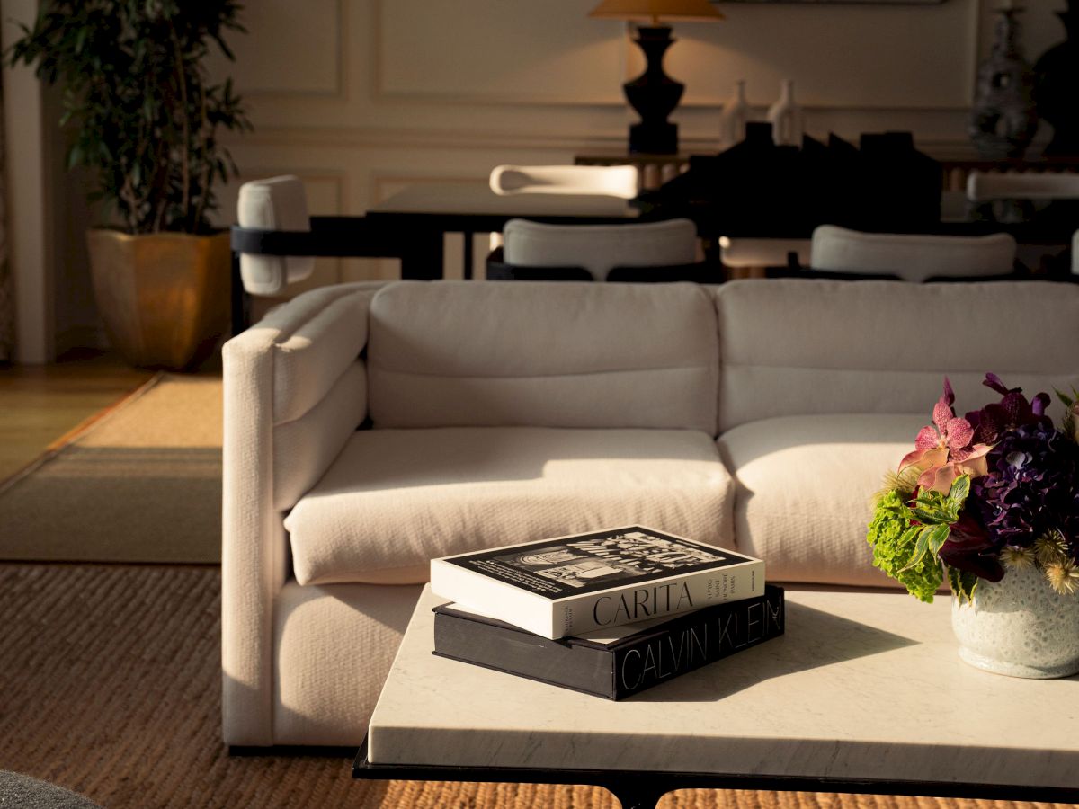 A cozy living room with a white sofa, books on a coffee table, a bouquet of flowers, and warm hanging lamps.