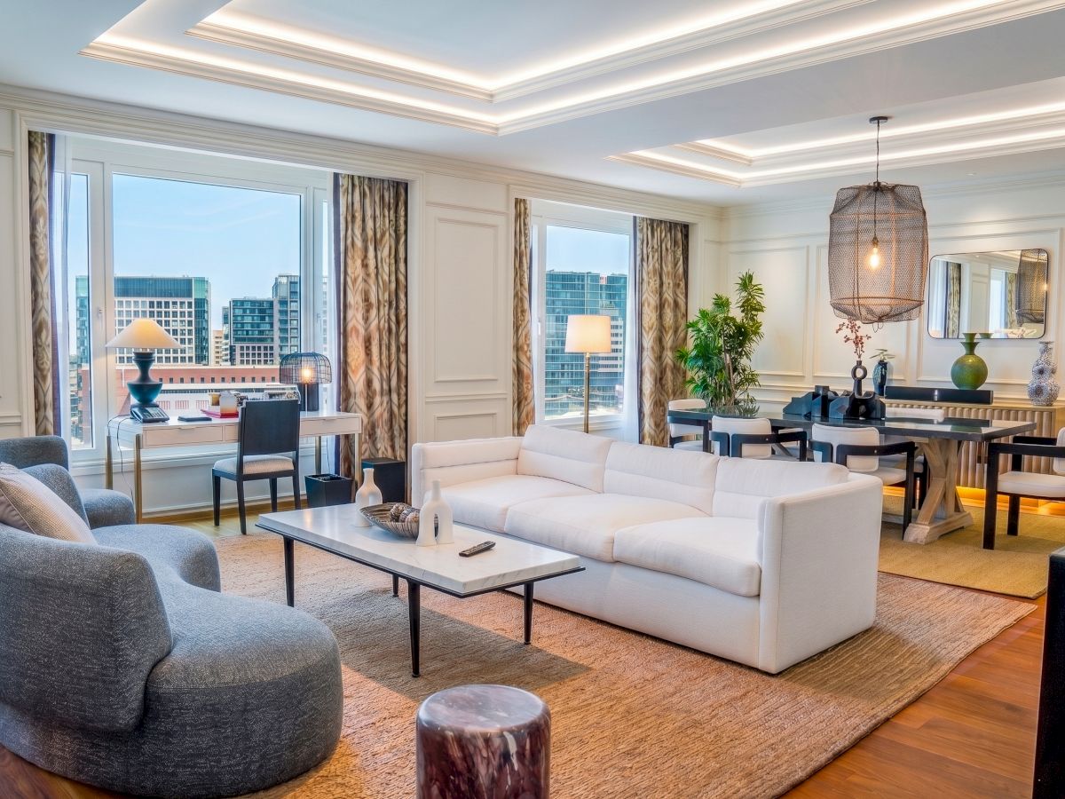 Modern living room with a white sectional, coffee table, pendant lights, and floor-to-ceiling windows facing a city skyline.