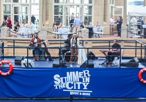 A band is performing on a stage with a sign that reads "Summer in the City". People are dining and socializing in the background.